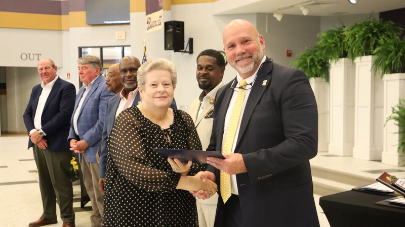 An award recipient poses with a presenter while holding a certificate at a recognition event, with attendees seated at tables in the background.