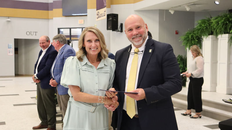 An award recipient poses with a presenter while holding a certificate at a recognition event, with attendees seated at tables in the background.