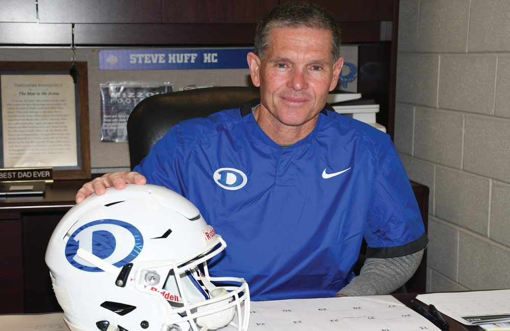 Photo of Coach Huff in his office wearing a blue Decatur shirt and holding a football helmet