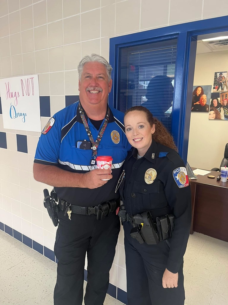 Two police officers, a man and a woman, stand side-by-side in a school hallway and smile for a photo. Both officers wear dark blue uniforms with patches on their sleeves that read "Police Decatur." The man on the left is tall with graying hair and a mustache; he wears a blue and black short-sleeed uniform shirt and holds a red and white coffee cup. The woman on the right has long, reddish-brown curly hair pulled back in a ponytail and wears a long-sleeved navy uniform.  They are standing in front of a blue doorway. To the left, a sign on the white-tiled wall reads "Hugs NOT Drugs" in blue and pink lettering. In the background, inside the office, photos are visible on the wall and a desk is partially visible.