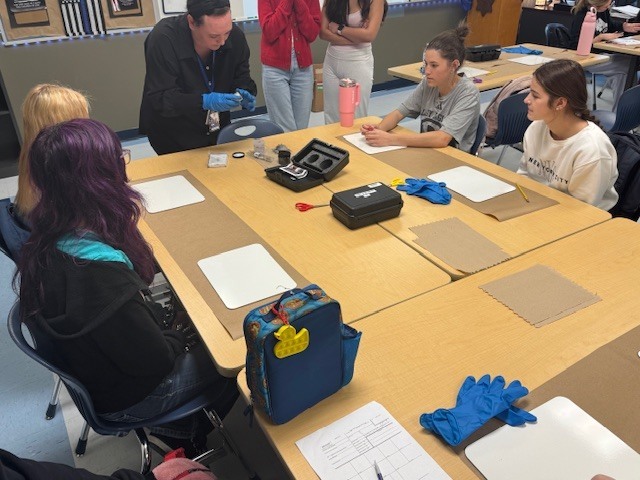 A close-up photograph capturing high school students actively engaged in a Law Enforcement I class fingerprinting lab. Three large wooden tables are joined together, covered by brown butcher paper work surfaces and white cards. In the foreground, a student's hand with black ink stains holds a small black cylindrical object (fingerprint powder container) over a fingerprint card. The card features several detailed black ink-rolled fingerprints in labeled boxes. Another student is visible in the upper background, seemingly about to ink their hands or work on another card. Multiple fingerprint cards, some with existing prints, pencils, and additional black lids are scattered across the table.