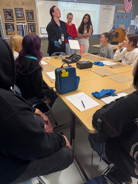 A close-up photograph capturing high school students actively engaged in a Law Enforcement I class fingerprinting lab. Three large wooden tables are joined together, covered by brown butcher paper work surfaces and white cards. In the foreground, a student's hand with black ink stains holds a small black cylindrical object (fingerprint powder container) over a fingerprint card. The card features several detailed black ink-rolled fingerprints in labeled boxes. Another student is visible in the upper background, seemingly about to ink their hands or work on another card. Multiple fingerprint cards, some with existing prints, pencils, and additional black lids are scattered across the table.