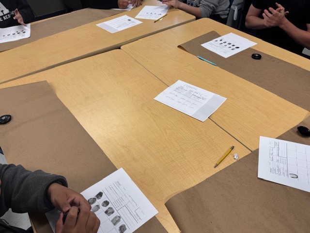 A close-up photograph capturing high school students actively engaged in a Law Enforcement I class fingerprinting lab. Three large wooden tables are joined together, covered by brown butcher paper work surfaces and white cards. In the foreground, a student's hand with black ink stains holds a small black cylindrical object (fingerprint powder container) over a fingerprint card. The card features several detailed black ink-rolled fingerprints in labeled boxes. Another student is visible in the upper background, seemingly about to ink their hands or work on another card. Multiple fingerprint cards, some with existing prints, pencils, and additional black lids are scattered across the table.