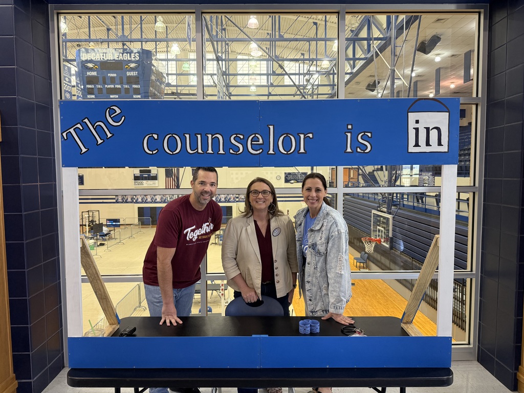 Three school counselors stand smiling behind a large, handmade blue and white wooden booth that reads "The counselor is in" with a bucket icon. They are positioned in a school hallway with a large window behind them overlooking the Decatur High School gymnasium and basketball court. The counselor on the left wears a maroon shirt, the middle counselor wears a tan blazer, and the counselor on the right wears a denim jacket.