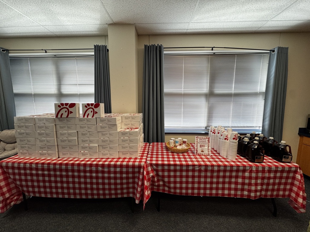 long table with a red and white checkered tablecloth is stacked with white boxes of Chick-fil-A meals, stacked high.  Next to the boxes, there are pitchers of drinks and stacks of white styrofoam cups.  A printed sign with the event details is also on the table.