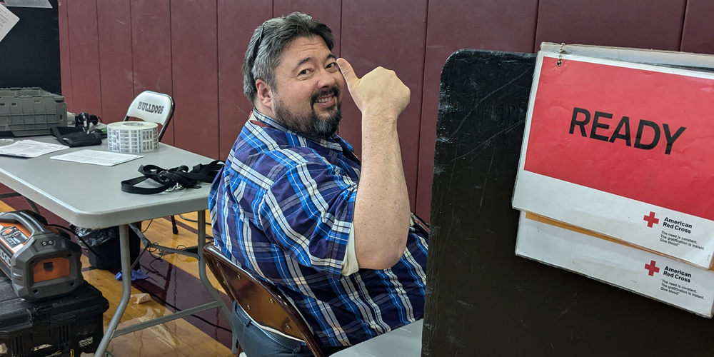 teacher gives thumbs up sign as he sits at blood drive next to "READY" sign.