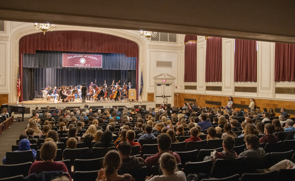 stage with orchestra and speaker; audience visible in auditorium