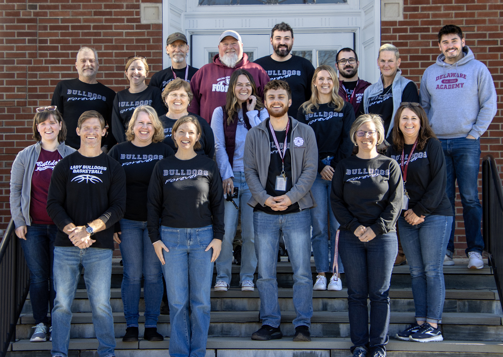 18 adults pose in front of school wearing Bulldgo shirts