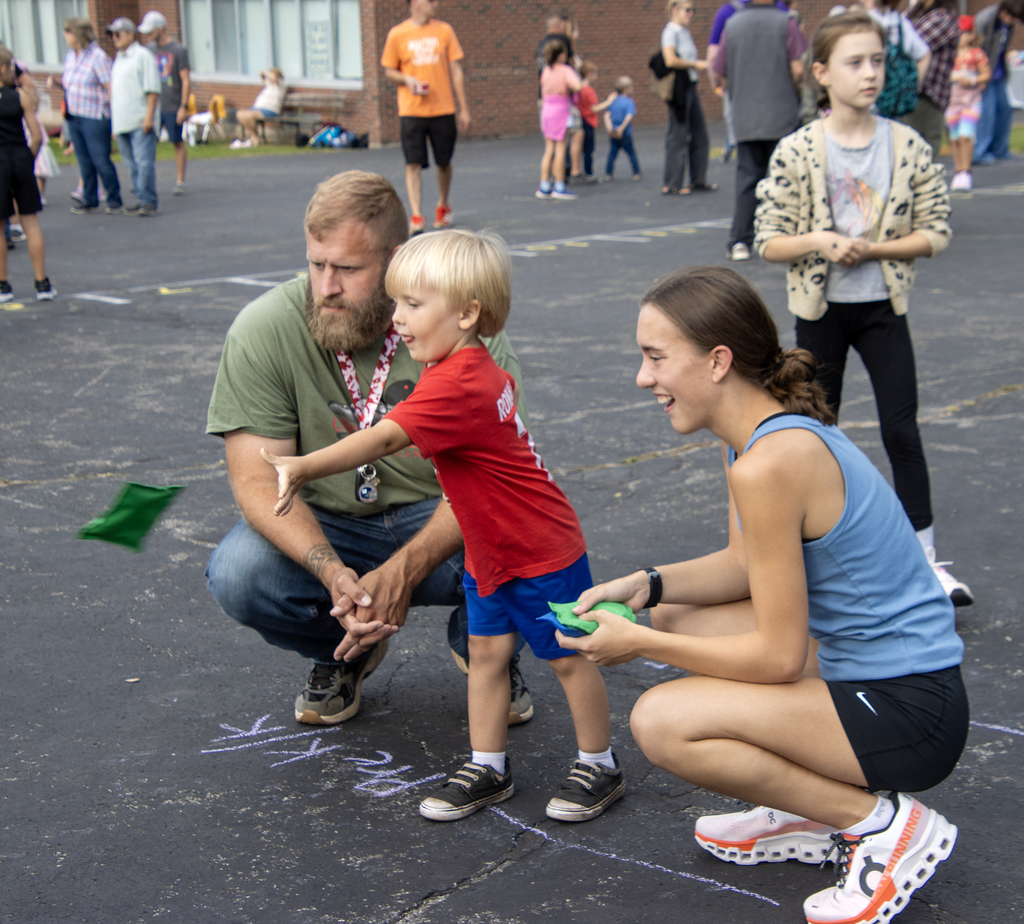 adult and older student kneel next to elementary student throwing a beanbaf