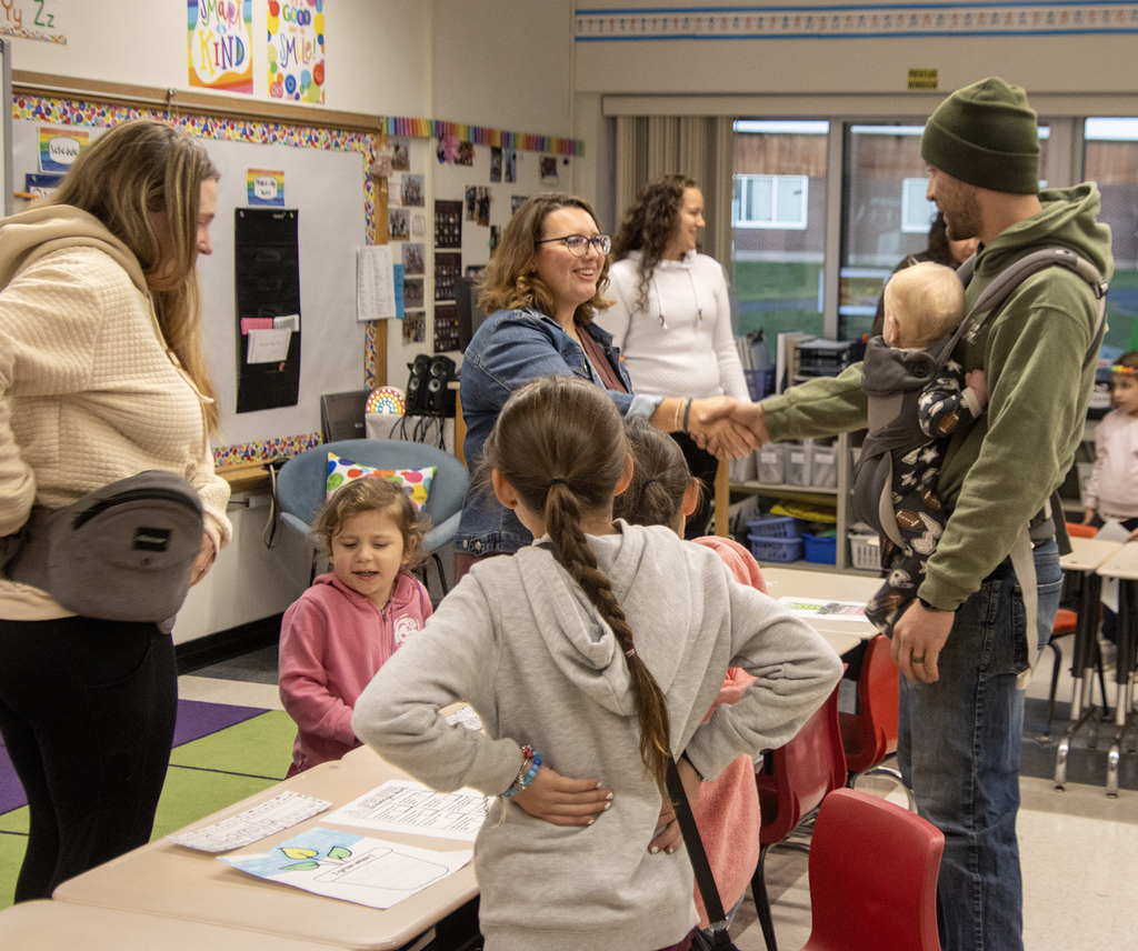 teacher shakes hands with parent holding a baby, other parent and 3 other children are nearby