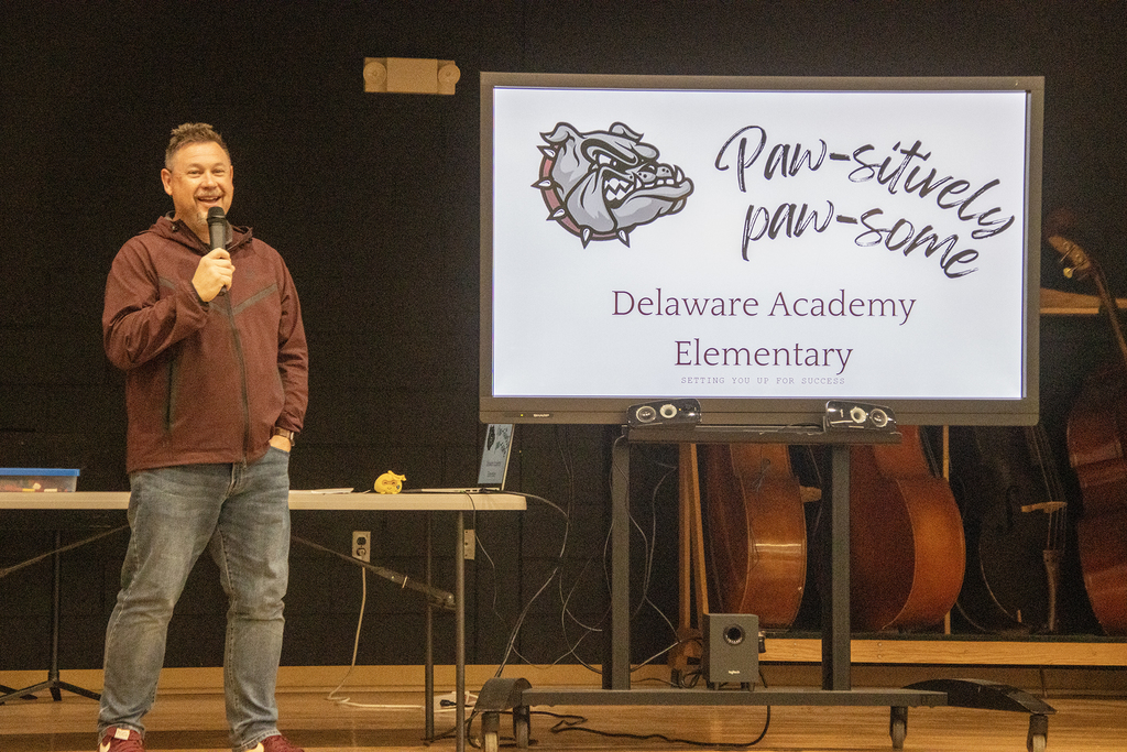 Mr. Albright smiles on stage next to sign reading Paw-sitively Paw-some! Delaware Academy Elementary