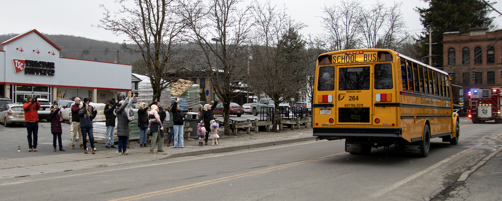 adults cheer the varsity girls bus on as it passes Tractor Supply