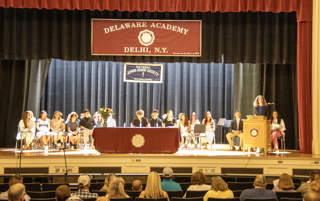 Student at lectern on stage; members and inductees of the NJHS are sitting on stage