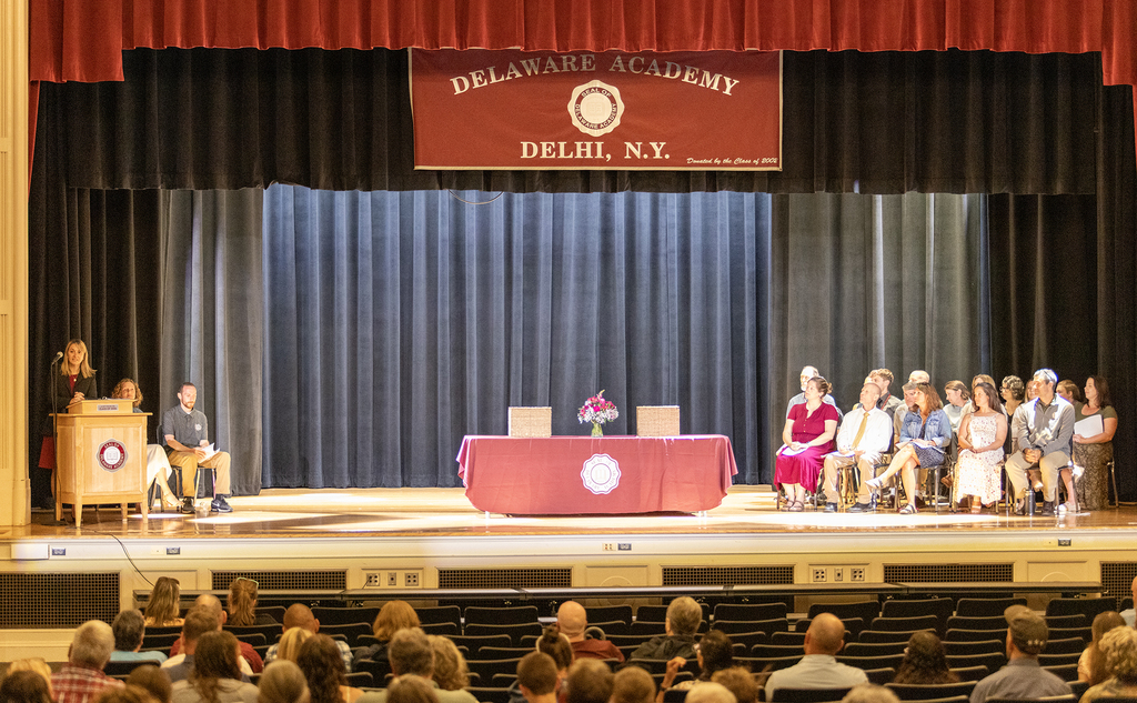 Ms. Trask speaking, Mrs. Kane and Mr. Chase seated behind her, teachers sitting on stage