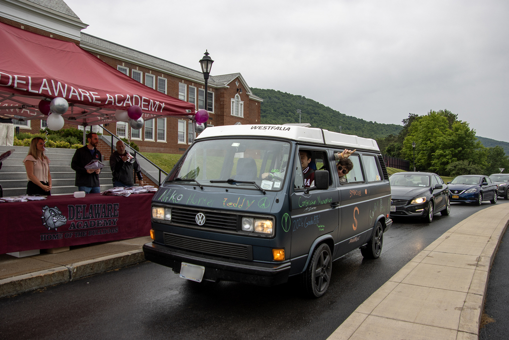 students in old VW van picking up their caps and gowns in front of school