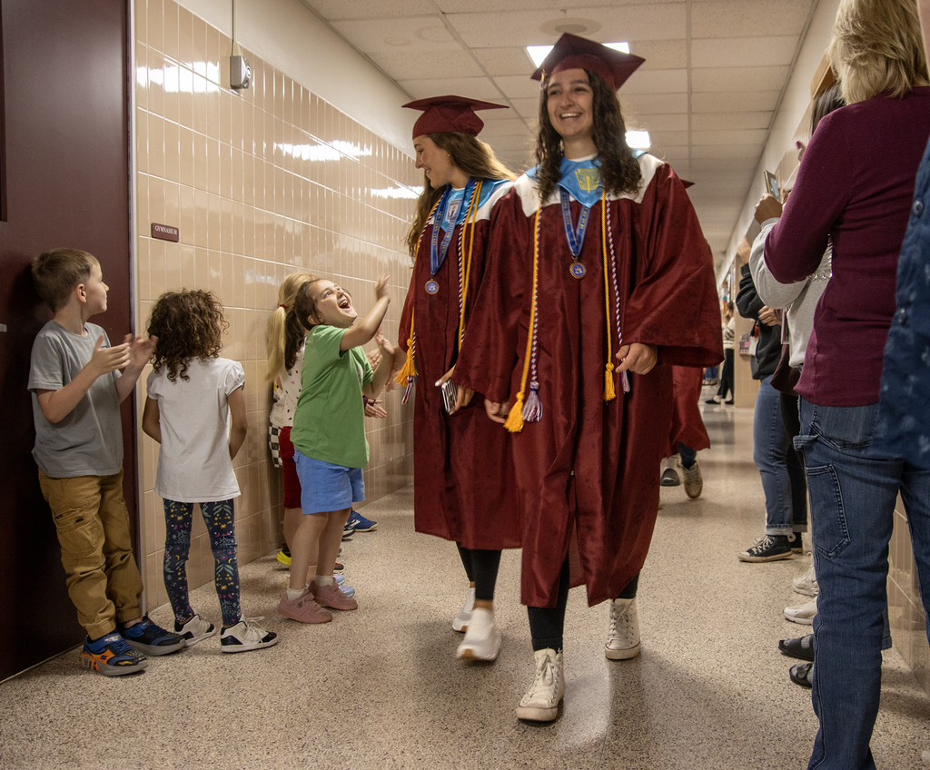seniors in caps and gowns walk through the hallway, students offering high fives