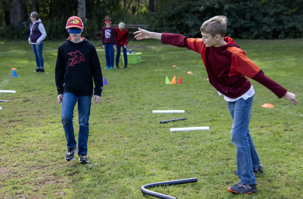 blindfolded student navigating an obstacle course, guided by another student, who is hilariously using his hands to direct the student