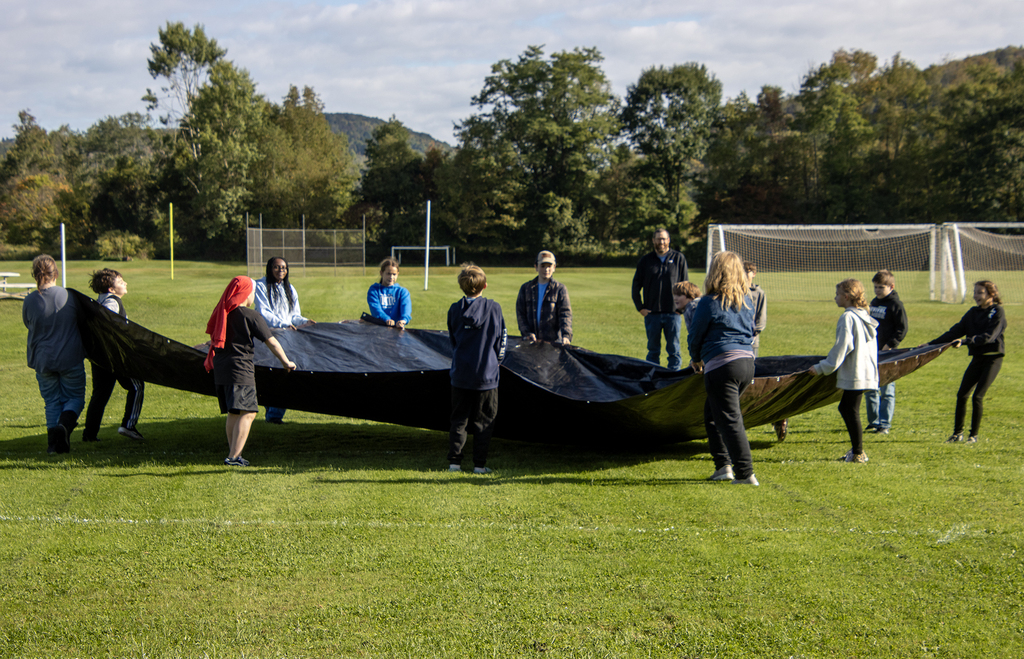 students holding a large piece of black plastic tarp, teacher watching