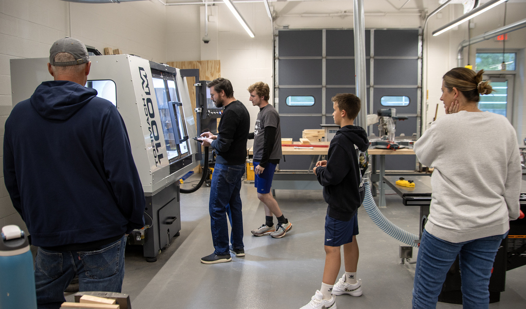 two students and two adults watch a teacher using a CNC milling machine in the tech lab
