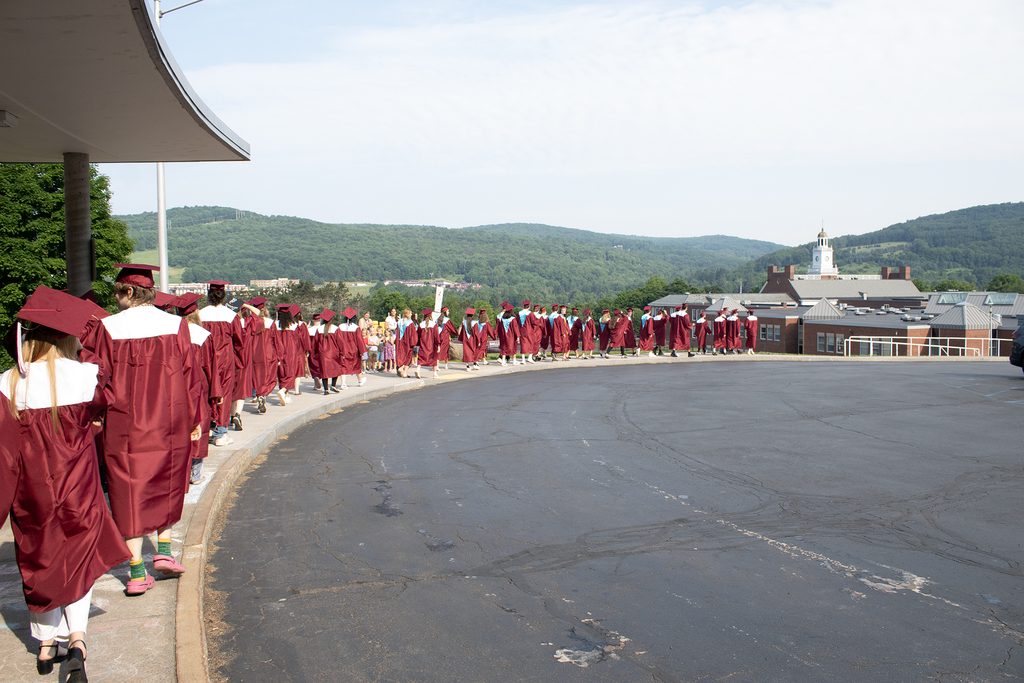 seniors in caps and gowns walking on sidewalk with school in background