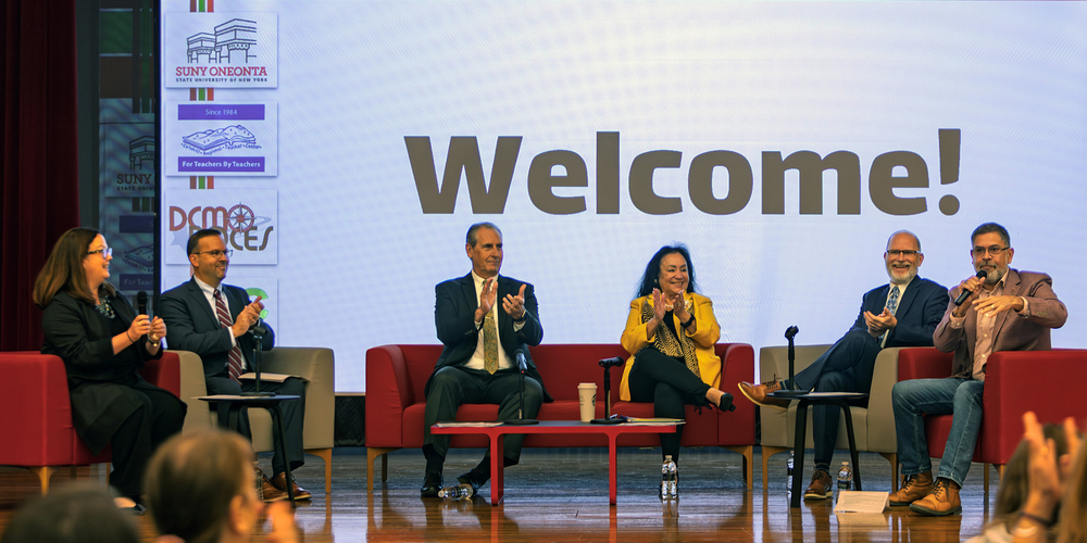 6 adults sit on stage, Welcome! is on screen behind them