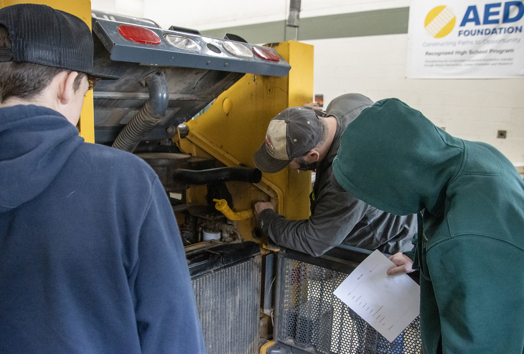 an insturctor shows 2 visiting students where somethin is on a skid steer