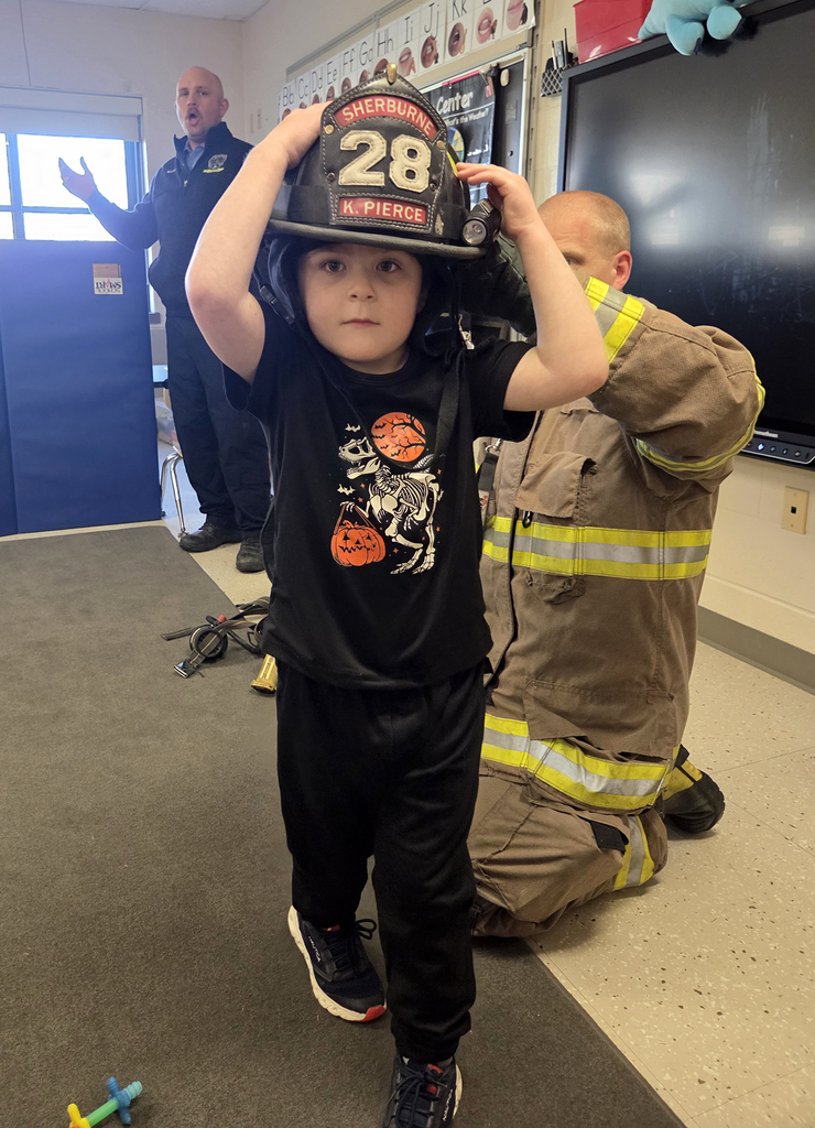 firefighter in turnout gear with students and another firefighter; student wearing firefighter's helmet