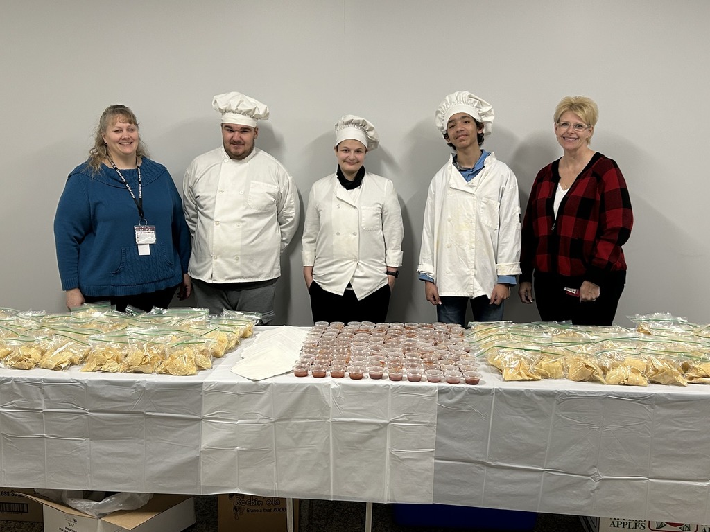 3 students in chefs uniforms stand with 2 adults behind table with chips and salsa