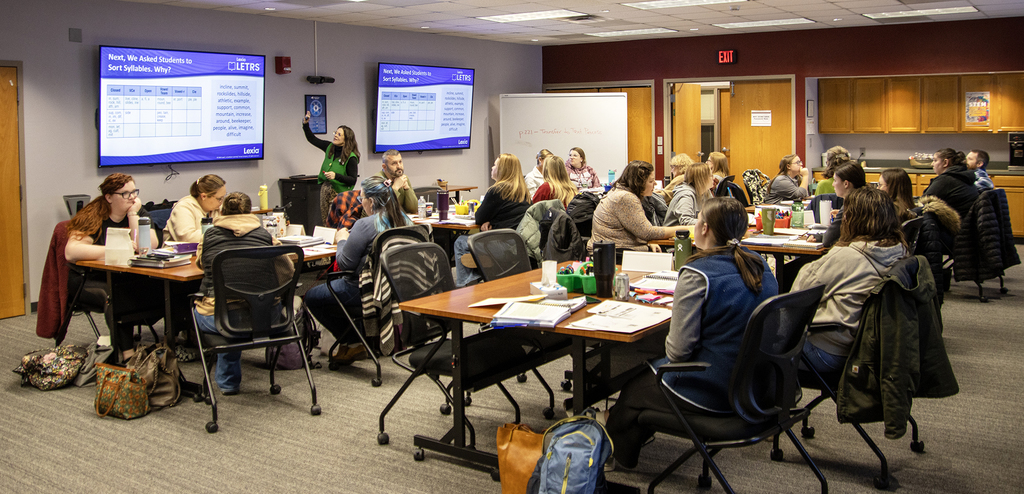 presenter gesturing to a screen in room full or workshop participants