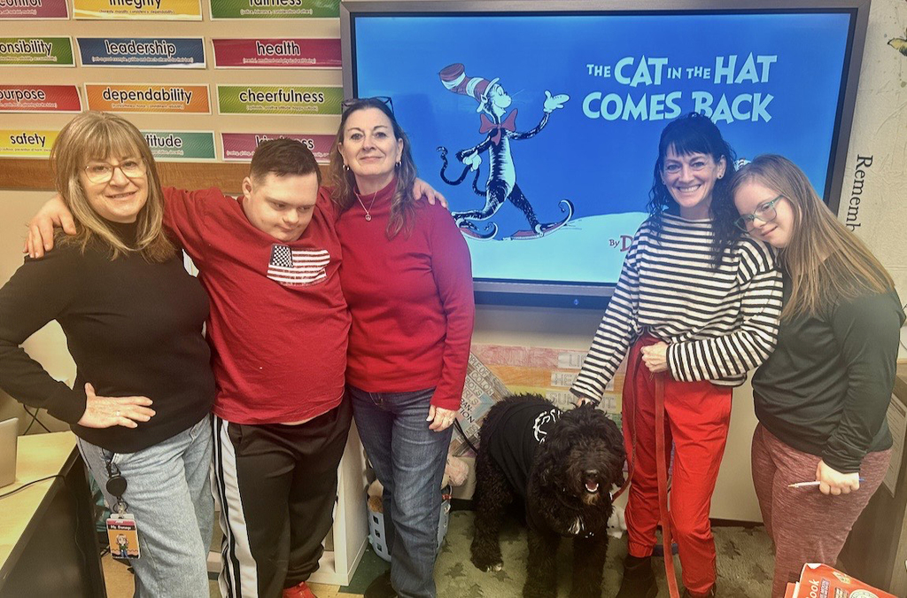 students and treachers pose with Heltie the dog; The Cat in the Hat Comes Back book is on the screen