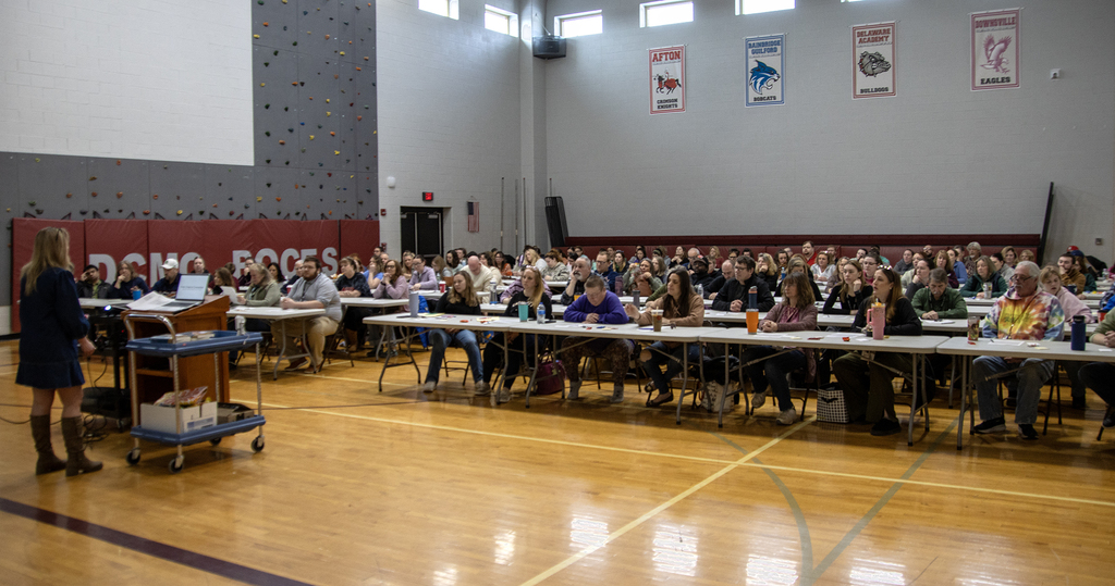Sara Stafford at lectern, audience sitting at long conference tables in gym