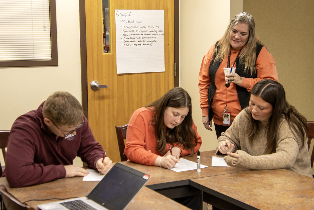 superintendent looks on as 3 students write at a table