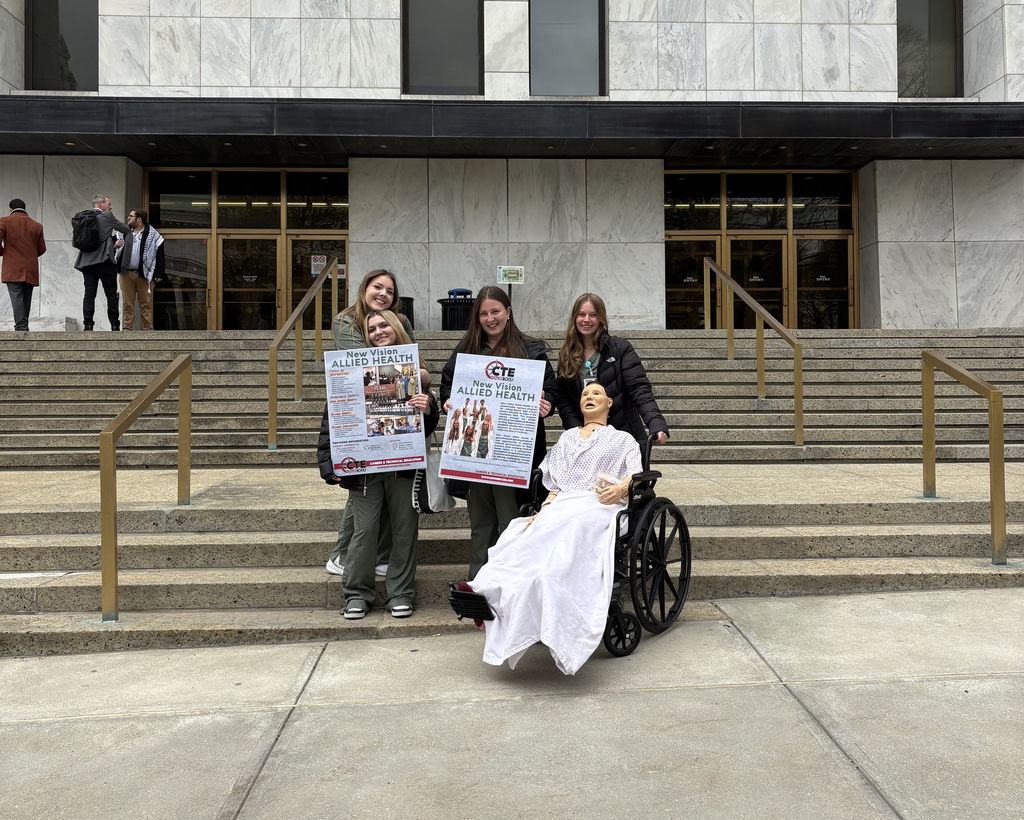 4 students pose with posters ands a medical mannequin in a wheelchair in front of state legislative office building