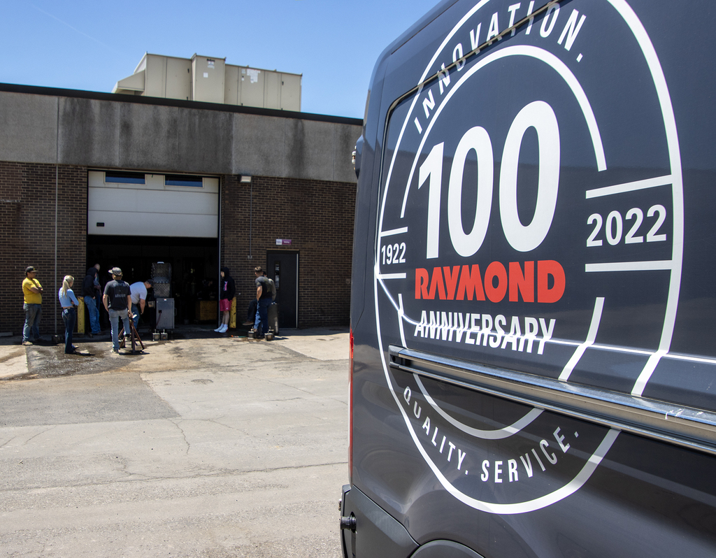 Foreground: Raymond van with anniversary logo/seal. Background: Students and teachers gathered around the pressure test machine