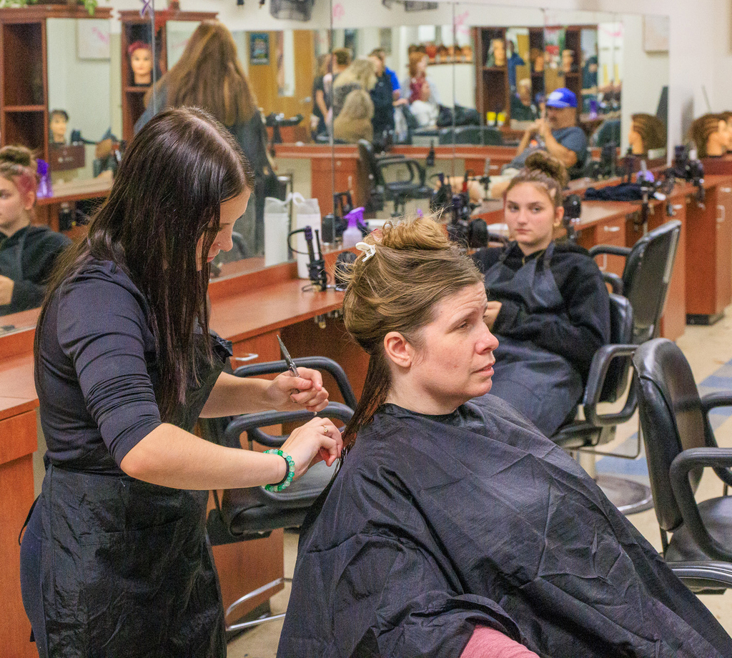 a cosmetology student is shown working on an adult's hair; another student is in a chair behind them