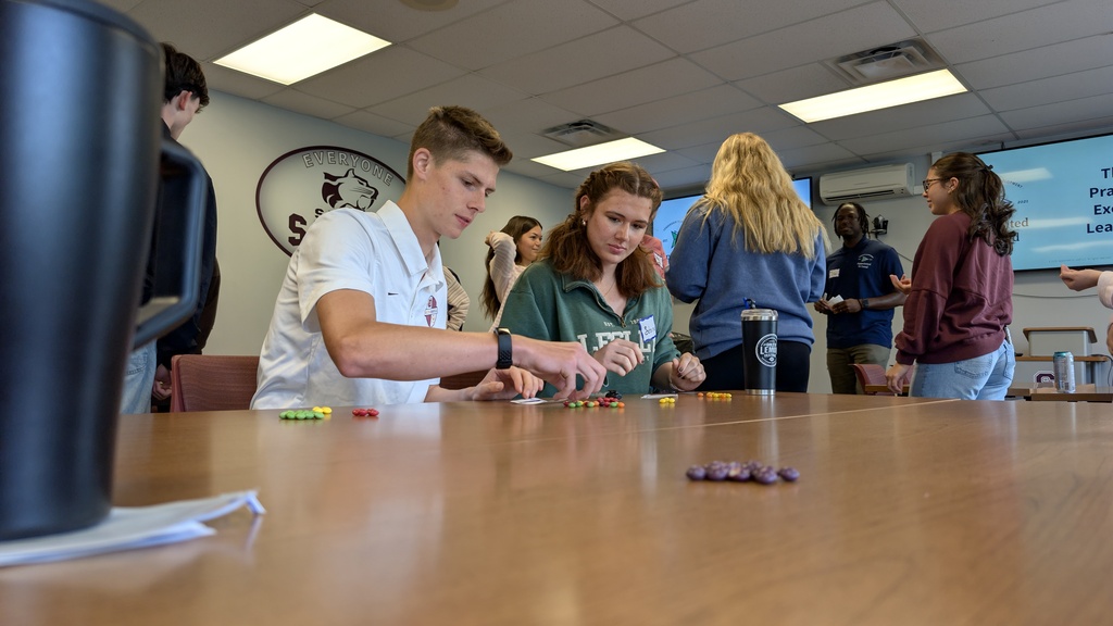 two students work at a challenge on a table while other students can be seen in the background
