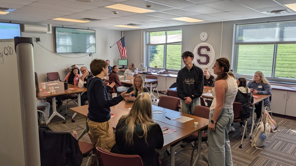 3 students stand as they talk across a table in a conference room
