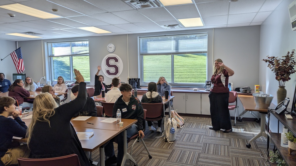 an adult presenter speaks to students sitting at tables in a conference room