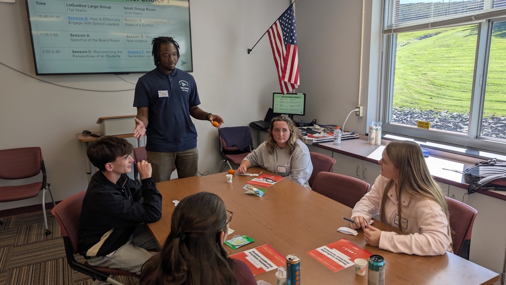 A standing presenter speaks to 4 students at a table in a classroom