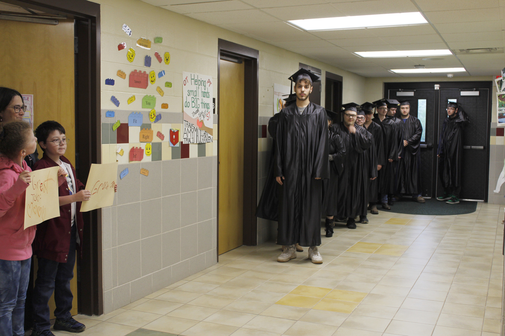 2 students holding signs with teacher look at seniors in hall