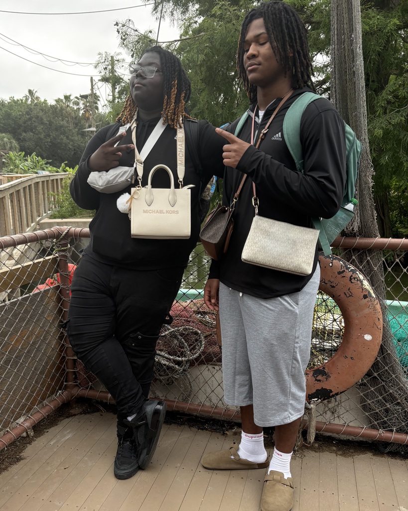 Two individuals on a wooden bridge. One has a backpack and points, the other wears a white bag. Behind them is a fence and trees.