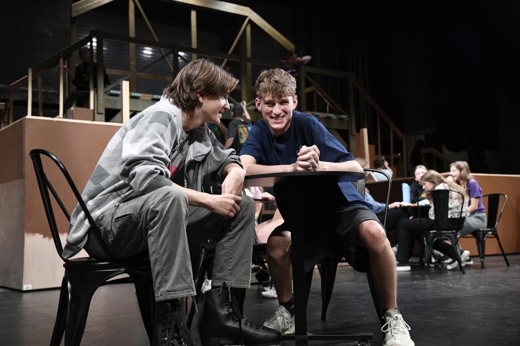 Two young men sit at a table, smiling, with an audience in the background. One wears a blue shirt and white shoes; the other wears a gray shirt and pants.