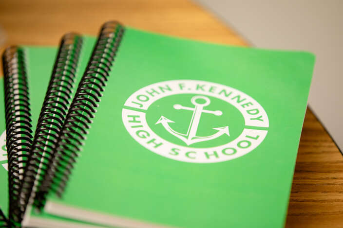 Photo of green notebooks stacked on top of a table. Top notebook has the logo of white John F. Kennedy High School logo