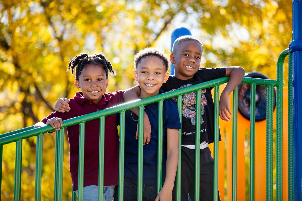 three boys smile, arm-in-arm, on a jungle gym with fall leaves in the background