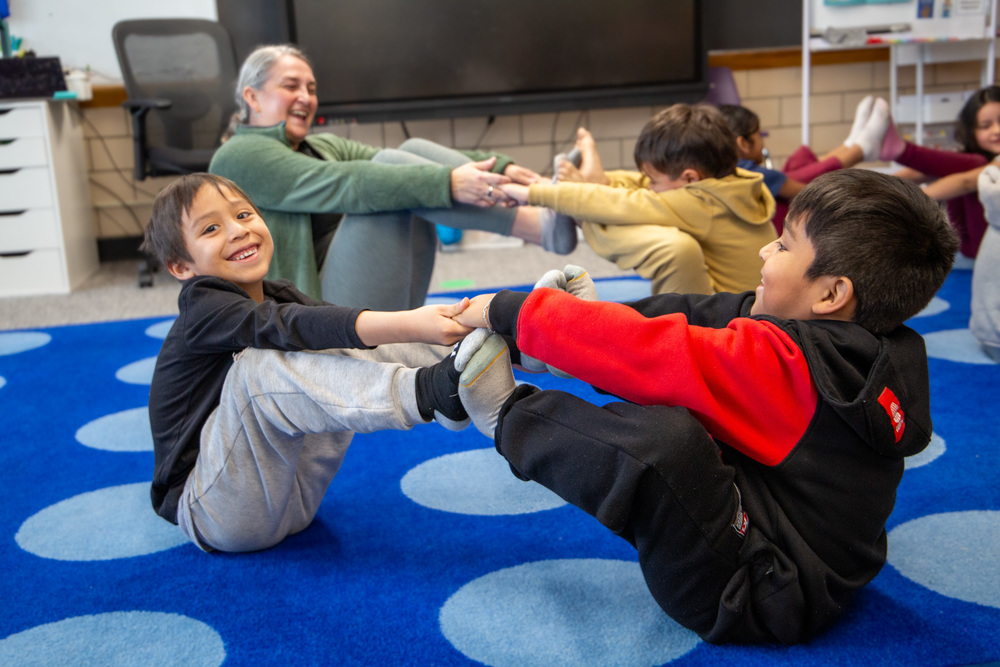 A student attempts a yoga pose with another student. They sit across from one another as their feet touch and they hold hands. 
