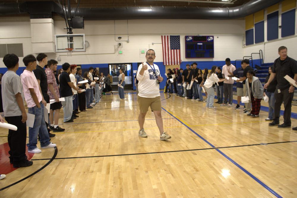 NEC staff member walking in the gym holding a thumbs up and smiling while two lines of students stand on either side of him