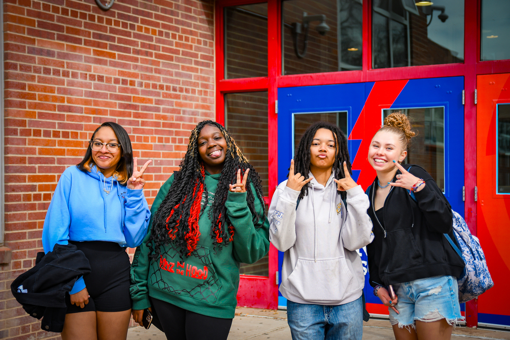 Manual students smile and hold up peace signs in front of the school