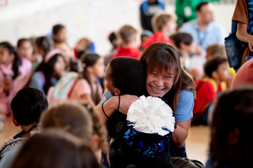 McMeen student gets a hug from a smiling teacher holding pompoms