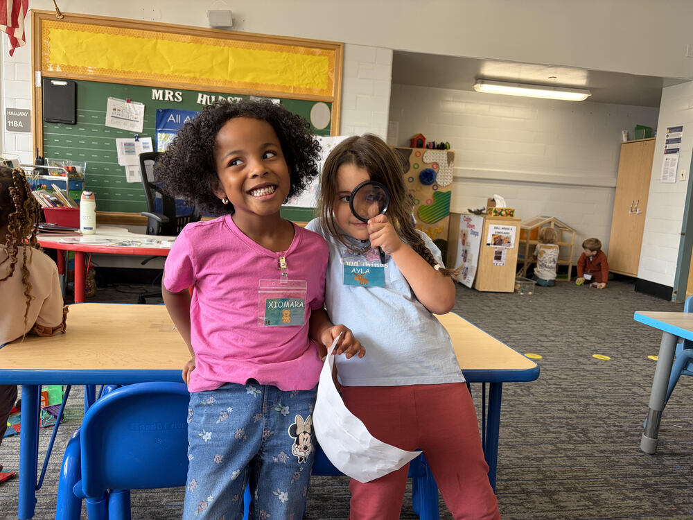 a three-year-old and four-year-old play together in a classroom with one girl holding a magnifying glass up to her eye