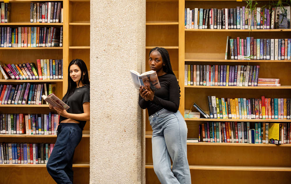 Two highschoolers holding books while leaning by a column in the library